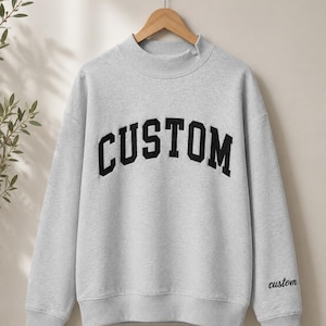 May include: A light gray crewneck sweatshirt featuring a mock neck collar and long sleeves. The word CUSTOM is printed in large, bold, black block letters across the chest, and the word custom is embroidered in a smaller black script on the left sleeve.
