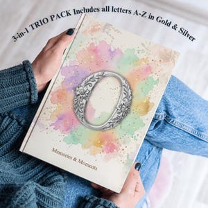 Op de afbeelding: Een rechthoekig boek met een witte kaft met een centrale zilveren florale letter O omringd door een pastelkleurig aquarelontwerp. De tekst 3-in-1 TRIO PACK Includes all letters A-Z in Gold & Silver en Memories & Moments is afgedrukt.