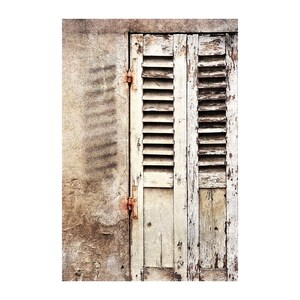 May include: A weathered wooden window shutter with white paint peeling off. The shutter is closed and has multiple horizontal slats. The shutter is attached to a wall with rusty hinges.