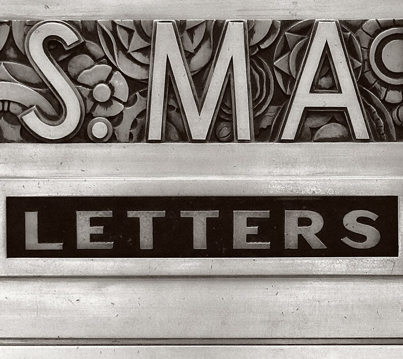 May include: A vintage silver mailbox with the word "LETTERS" in raised lettering on a black background and the word "SMA" in raised lettering on a decorative background.