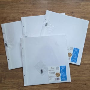 May include: A set of four square white photo albums with a three-ring binder mechanism. Each album features a clear plastic pouch on the front containing small black metal screws for assembly. The covers are plain white with a blue and white label.