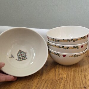 May include: A set of four cream-colored ceramic bowls. One bowl is shown individually, featuring a small illustration of a blue birdhouse with red hearts and a bird. The other three bowls are stacked, decorated with a vine pattern and red hearts.
