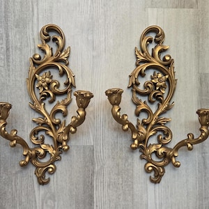 May include: A pair of gold-colored wall sconces featuring intricate scrollwork, floral motifs, and a single candle holder arm on each piece. The decorative wall-mounted fixtures display a symmetrical design with ornate, swirling leaf patterns.