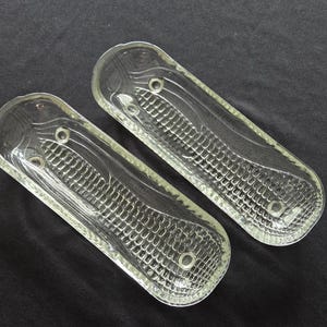May include: Two clear glass dishes, each shaped like an elongated oval with rounded ends. The interior surface of each dish features a textured, embossed pattern resembling a corn cob, complete with kernels and husks, and two small circular indentations.