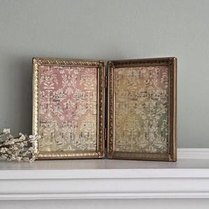 May include: A hinged double picture frame with a metallic gold finish and ornate edges. The two panels contain a decorative paper insert featuring a pink and yellow damask pattern layered over vintage musical sheet music notation.