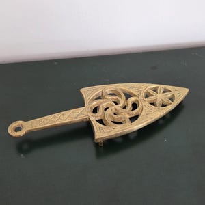 May include: A gold-toned metal trivet featuring a triangular shape with a long handle. The surface displays an intricate openwork design with a central swirling pattern and a six-petaled flower motif near the pointed end, finished with etched details.