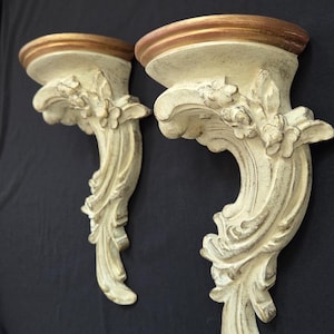 May include: A pair of wall-mounted shelves featuring an off-white finish with distressed gold accents. Each shelf has a semi-circular top platform and a curved, ornate support bracket decorated with carved floral and scrolling leaf motifs.