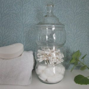 May include: A clear glass apothecary jar with a lid, stacked with cotton swabs in the middle section and white cotton rounds in the bottom section. Beside the jar sits a folded white towel topped with a single white bar of soap.
