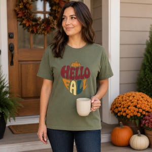 May include: An olive green short-sleeve t-shirt featuring a graphic print of a pumpkin shape containing the text "HELLO FALL" in stacked, bold, multi-colored lettering. The shirt is made of a solid fabric and has a crew neckline.