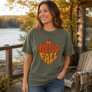 May include: An olive green short-sleeve t-shirt featuring a graphic print of an orange pumpkin above the text "HELLO FALL" in a stacked, retro-inspired font. The shirt has a crew neckline and a relaxed fit, showcasing a solid, muted color.