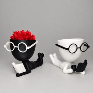 May include: Two 3D-printed planters shaped like figures lying on their stomachs reading books. One planter is black with white glasses and a white book, holding red flowers. The other is white with black glasses and a black book.