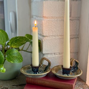 Peut inclure: Deux bougies effilées blanc cassé allumées dans des bougeoirs en céramique faits main avec poignées. Les bougeoirs présentent un vernis brillant aux tons bleus et beiges. Les bougies sont posées sur deux livres rouges empilés avec lettrage doré.