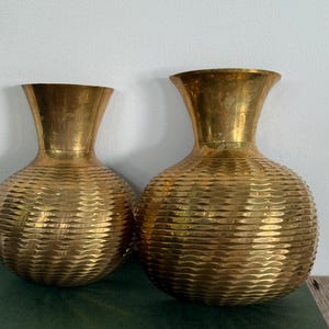Peut inclure: Deux vases en laiton poli avec un corps arrondi et bulbeux et un col évasé. La surface de chaque vase présente un motif horizontal texturé en forme de vagues qui fait le tour de toute la circonférence de la base arrondie.