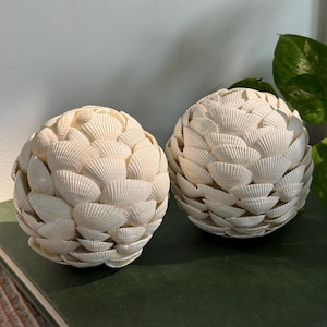 May include: Two spherical decorative objects covered in overlapping white scallop shells. Each orb features a dense, layered texture created by the arrangement of the individual shells, resulting in a rounded, textured surface with a uniform off-white color.