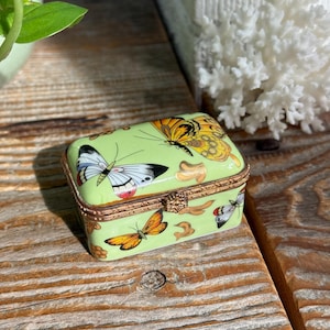 May include: A rectangular light green trinket box with a hinged lid, featuring colourful butterfly illustrations in orange, yellow, black, and white, accented with gold-toned metal trim and a small decorative clasp on the front edge.