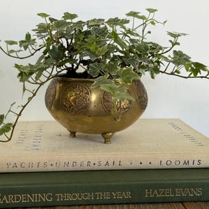 Peut inclure: Un bol en laiton à trois petits pieds avec des panneaux floraux circulaires en relief, contenant une plante de lierre vert et blanc. Le bol repose sur deux livres empilés, un beige intitulé Yachts Under Sail et un vert Gardening Through the Year.