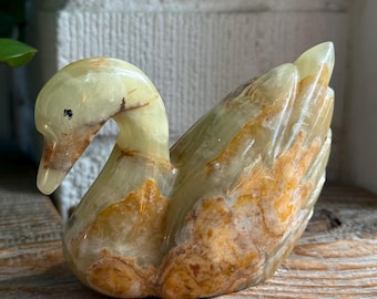 May include: A carved stone swan figurine featuring a smooth, polished surface with translucent pale green and opaque amber-colored mineral veining throughout the body, neck, and head, with a small dark spot marking the eye area.