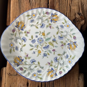 May include: A round ceramic plate with a scalloped edge and a thin blue rim. The surface features a repeating pattern of stylised floral motifs in shades of yellow, blue, purple, and green on a white background, arranged in a flowing design.