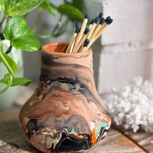 Peut inclure: Un petit vase en céramique arrondi présentant un motif marbré avec des tourbillons de brun, noir, orange et bleu sarcelle. Le vase possède une base large et une ouverture étroite, contenant un groupe d'allumettes en bois à bouts noirs.