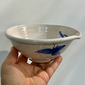 May include: A speckled off-white ceramic bowl with a glossy finish, featuring a hand-painted blue floral design on the exterior and a small pouring spout on the rim. The bowl has a wide, circular opening and a tapered base with a smooth texture.