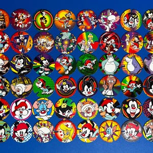 May include: A collection of sixty circular cardboard pogs featuring colorful illustrations of characters from Animaniacs and Pinky and the Brain. Each disc displays unique cartoon imagery, including various expressions and scenes set against vibrant backgrounds.