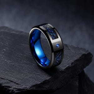 May include: A black band ring featuring a polished finish with a central inlay of blue and black textured material. The ring has a bright blue interior and a single small round blue gemstone set into the outer surface of the band.