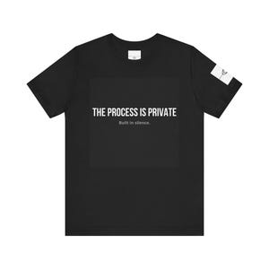 May include: A black short-sleeve t-shirt featuring the white text THE PROCESS IS PRIVATE and Built in silence. centered on the chest. A small white rectangular patch with a black line drawing of a flower is located on the left sleeve.