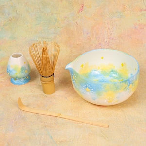 May include: A matcha tea set featuring a ceramic bowl and a whisk holder with a blue and yellow star pattern, a bamboo whisk, and a bamboo tea scoop. The bowl has a pouring spout and a smooth interior finish, while the whisk has thin bristles.