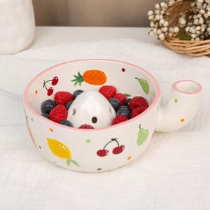 Pode incluir: Um espremedor de frutas de cerâmica branca com borda rosa e alça. O exterior e o interior apresentam ilustrações coloridas pintadas de cerejas, abacaxis, limões e peras. Um cone central branco com orifícios fica dentro da tigela.
