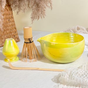 May include: A matcha tea set featuring a bright yellow ceramic bowl with a pouring spout, a matching yellow ceramic whisk holder, a bamboo whisk with thin tines, and a long, curved bamboo tea scoop, all arranged together for tea preparation.