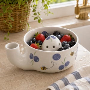 May include: A white ceramic berry bowl with a side spout and a central drainage dome. The exterior features hand-painted blue blueberries and small yellow and black bees. The bowl is filled with fresh strawberries, blueberries, and blackberries.