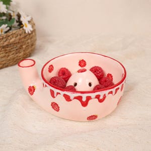 May include: A light pink ceramic yarn bowl featuring a painted red raspberry pattern and a white drip glaze design along the rim. The bowl includes a side notch for yarn and a central post with two small holes, currently filled with fresh red raspberries.