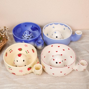 May include: Four ceramic yarn bowls with side yarn guides and central domes. One is blue with whale motifs, one is light blue with floral patterns, one is cream with strawberry and cherry prints, and one is cream with small red heart patterns.