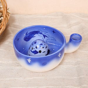 May include: A blue and white ceramic yarn bowl with a smooth gradient finish. The interior features a whale shark illustration, small fish, and stars. A central dome with holes and a side notch allow for yarn to pass through during knitting projects.