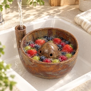 May include: A round, brown, speckled ceramic berry colander with a tall side spout and a central dome featuring drainage holes. The bowl is filled with fresh strawberries, blueberries, and green grapes being rinsed under a stream of water.