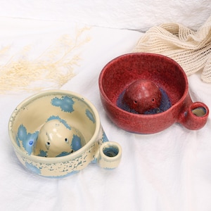 May include: Two ceramic shaving bowls with a central raised dome and a side handle. One bowl is cream-colored with blue speckled patterns, while the other is a deep red with a textured finish and a dark blue base around the central dome.