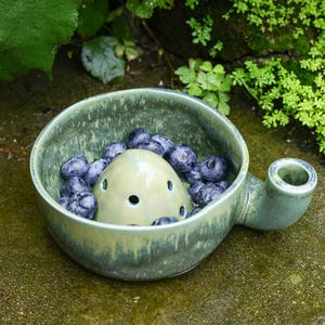 May include: A sage green ceramic berry bowl with a mottled glaze finish and a central dome featuring small drainage holes. The bowl includes a side handle with a hollow opening and is filled with a cluster of fresh, dark blue round blueberries.