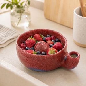 May include: A round, speckled red ceramic bowl with a small, cylindrical side handle, filled with a mixture of fresh strawberries, raspberries, and blueberries. The bowl has a textured, mottled finish with hints of blue near the base.
