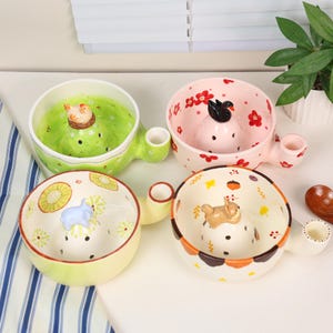 May include: Four ceramic bowls with handles, each featuring a central raised mound with a small animal figurine on top and perforated holes. The bowls have distinct designs: green with a chicken, pink with a black swan, yellow with a blue animal, and cream with a brown animal, all decorated with colourful patterns.