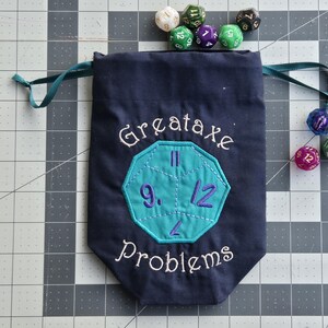 May include: A dark blue fabric drawstring bag featuring an embroidered teal dodecahedron shape with purple numbers and the white stitched text "Greataxe Problems" above and below the shape, surrounded by several multi-colored polyhedral dice.