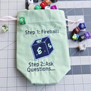 Könnte beinhalten: Ein hellgrüner Beutel mit Kordelzug und dunkelgrüner Stickerei mit der Aufschrift Step 1: Fireball! und Step 2: Ask Questions... sowie einer zentralen dunkelblauen Würfelgrafik. Der Beutel ist von mehreren bunten polyedrischen Würfeln umgeben.