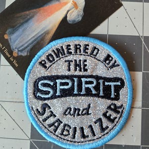 May include: A circular embroidered patch with a silver glitter background and a light blue border. The patch features black embroidered text that reads POWERED BY THE SPIRIT and STABILIZER, with the word SPIRIT in a larger font size.