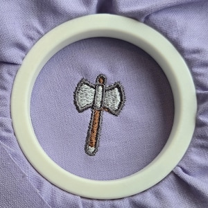 May include: A double-headed axe embroidery design stitched onto light purple fabric, secured within a white circular embroidery hoop. The axe features a silver blade with dark grey outlines and a brown handle with a silver base.