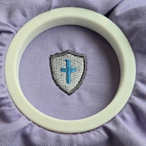 May include: A light purple fabric background featuring a centered embroidered shield emblem. The shield is outlined in dark gray, filled with light gray thread, and contains a bright blue cross in the center. A smooth white circular frame surrounds it.