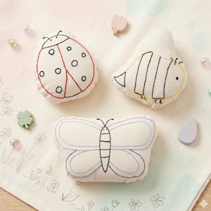 May include: Three small, stuffed fabric ornaments featuring embroidered insect designs: a ladybug with red and black stitching, a bee with yellow and black stitching, and a butterfly with purple and blue stitching on an off-white canvas.