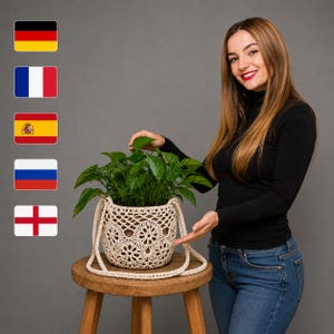 May include: A cream-colored, hand-crocheted plant pot cover featuring a circular floral pattern and two matching side handles. The cylindrical textile basket holds a lush green leafy plant and is displayed on a wooden stool.