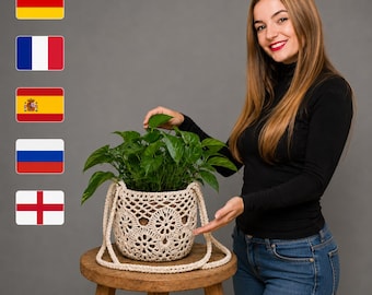 May include: A cream-colored, hand-crocheted plant pot cover featuring a circular floral pattern and two matching side handles. The cylindrical textile basket holds a lush green leafy plant and is displayed on a wooden stool.
