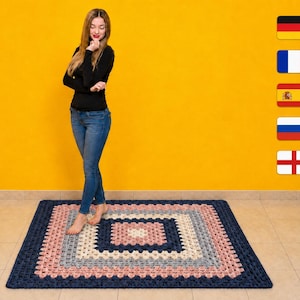 Könnte beinhalten: Ein quadratischer Häkelteppich mit einem konzentrischen Granny-Square-Muster in abwechselnden Streifen aus dunkelmarineblauem, hellgrauem, cremefarbenem und altrosa Garn. Der Teppich ist in einem strukturierten Lochmuster gefertigt und liegt flach.