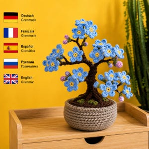 May include: A crocheted bonsai tree featuring light blue flowers with yellow centers and small pink buds. The tree has a dark brown trunk and branches, and it is potted in a round, light brown crocheted container with a textured surface.