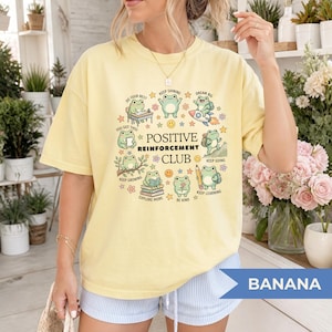 Może przedstawiać: Bladożółty t-shirt z okrągłą grafiką z napisem "POSITIVE REINFORCEMENT CLUB" otoczony ilustracjami zielonych żab wykonujących różne czynności, takie jak czytanie, wędrówki i jazda na rakiecie, z dodatkiem gwiazdek.